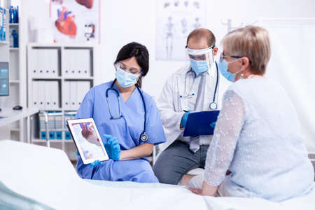 Senior woman during consultation at cardiologist discussing about heart disease wearing protection mask against coronavirus. Medical examination for infections, disease and diagnosis.の写真素材