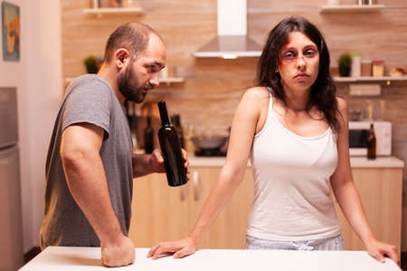 Agressive drunk husband scaring wife having bruises on entire face. Violent aggressive man abusing injuring terrified helpless, vulnerable, afraid, beaten and panicked woman.の写真素材