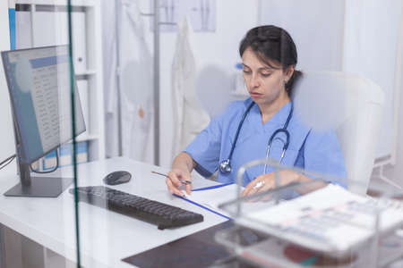 Medical staff writing notes on clipboard sitting at desk in hospital office. Healthcare physician specialist in medicine providing health care services treatment examination.の写真素材