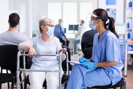 Nurse explaining diagnosis to disabled senior woman with walking frame wearing visor against infection with coronavirus in hospital hallway. Patient and medical staff in waiting area.の写真素材