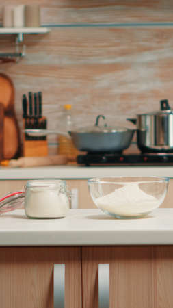 Pastry ingredients for homemade cakes and bread in empty kitchen. Modern dining room equipped with utensils ready for cooking with wheat flour in glass bowl and fresh eggs on tableの写真素材