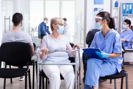 Senior woman with walking frame in hospital waiting room with nurse wearing face mask against coronaviurs contamination discussing illness treatment. Patient and medical staff in waiting area.の写真素材