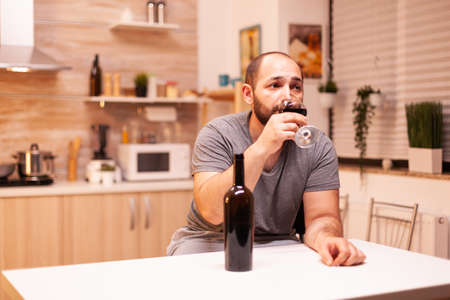 Upset young man drinking alone a bottle of red wine sitting at table in kitchen. Unhappy person disease and anxiety feeling exhausted with having alcoholism problems.の写真素材