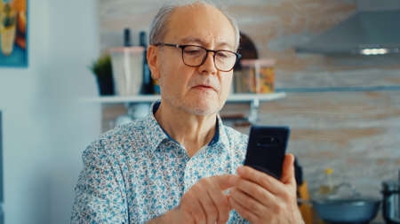 Old man surfing on social media using smartphone during breakfast in kitchen. Authentic portrait of retired senior enjoying modern internet online technologyの写真素材