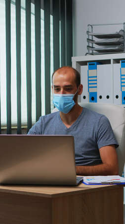 Man with mask having online meeting conference in modern office using laptop. Freelancer working in new normal office workplace chatting talking during virtual webinar, using internet technologyの写真素材