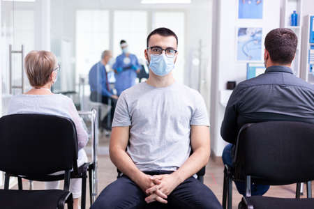 Portrait of tired young man with face mask against coronavirus in hospital waiting area looking at camera. Senior man during consultation in examination room.の写真素材