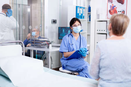 Medical staff wearing protective mask against coronavirus pandemic and talking with senior patient during consultation in medical examination room. Practitioner physician appointmentの写真素材
