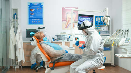 Stomatologist in coverall explaining proper dental hygiene using teeth skeleton during global pandemic. Medical team talking with woman wearing face shield, protection suit, mask and glovesの写真素材