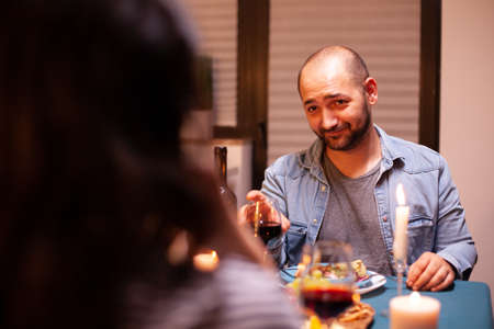 Man looking at wife while having romantic dinner and holding glass with red wine. Talking happy sitting at table dining room, enjoying the meal at home having romantic time at candle lights.の写真素材