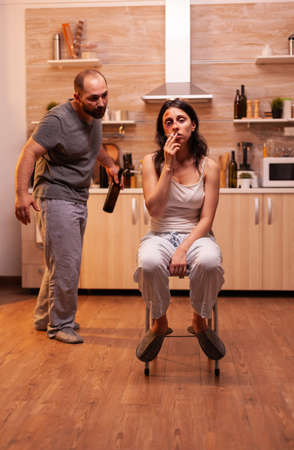 Abused woman smoking sitting in chair after being traumatised by drunk husband in kitchen. Violent aggressive man abusing injuring terrified helpless, vulnerable, afraid, beaten and panicked woman.の写真素材