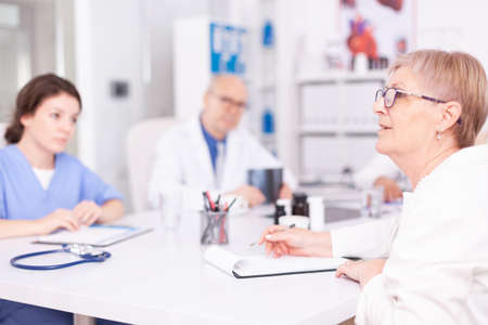 Mature woman doctor explaining diagnosis to medical staff in hospital conference room and having a discussion. Clinic expert therapist talking with colleagues about disease, medicine professionalの写真素材