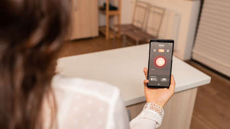 Woman in home kitchen using phone app to remotely control lighting system. Person in apartment...