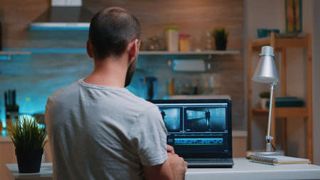 Colorist working remotely overtime from home using editing software sitting in front of laptop late at night. Videographer processing audio film montage on professional in modern kitchen in midnightの写真素材