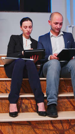Teamworkers sitting on stairs in corporate building doing overtime during deadline financial project looking on tablet and documents. Entrepreneurs working late together at corporate job.の写真素材