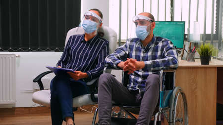 Businessman in wheelchair and woman partner with face masks having videomeeting in business office...