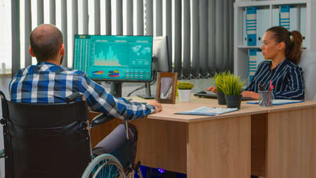 Disabled entrepreneur sitting in wheelchair immobilized analysing financial economy statistics on computer in business office discussing with colleague. Handicapped businessman using modern technologyの写真素材