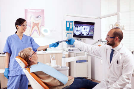 Dentist assistant giving doctor radiography of senior woman waiting. Medical teeth care taker holding patient radiography sitting on chair during consultation.の写真素材