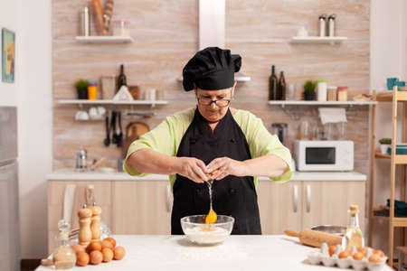 Elderly woman cracking eggs over wheat flour in home kitchen. Elderly pastry chef cracking egg on glass bowl for cake recipe in kitchen, mixing by hand, kneadingの写真素材