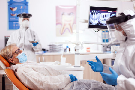 Dentist wearing safety gear against coronavirus talking about teeth treatment. Elderly woman in protective uniform during medical examination in dental clinic.の写真素材