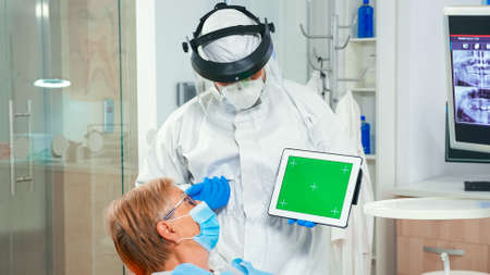Dentist in coverall pointing at green screen display during coronavirus epidemic. Explaining using...