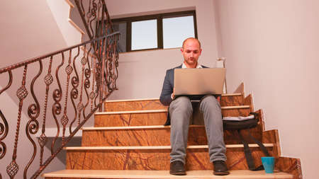Businessman typing on laptop speaking on phone sitting on stairs in finance corporate company...