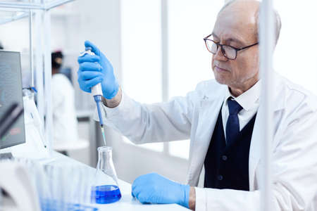 Elderly scientist doing genetic experiment in busy laboratory using sterile dropper with blue...