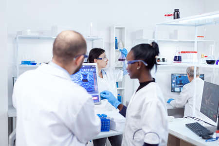Scientist holding glass flask with blue genetic material in busy laboratory. Multiethnic team of medical researchers working together in sterile lab wearing protection glasses and gloves.の写真素材