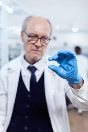 Concentrated senior scientist looking at glass slide with sample wearing protection gloves. Elderly researcher in sterile lab looking on microscope slide wearing lab coat.の写真素材