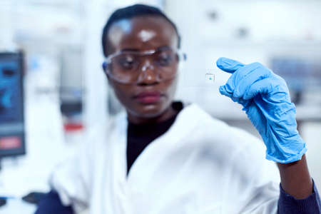 Multiethnic researcher during study holding microscope sample on glass slide. African healthcare scientist in biochemistry laboratory wearing sterile equipment.の写真素材