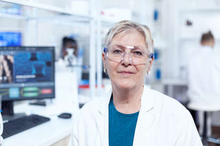 Close up portrait of biologist looking at camera in sterile laboratory. Elderly scientist wearing...