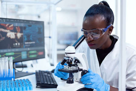 Medical researcher with african ethnicity using microscope doing investigation and evaluates sample. Black healthcare scientist in biochemistry laboratory wearing sterile equipment.の写真素材