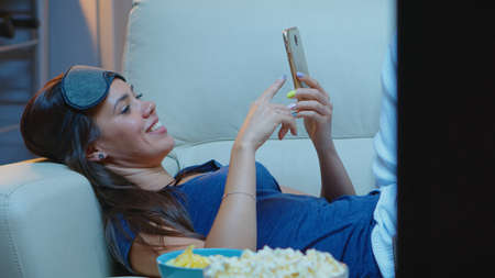 Lady with sleep mask on forehead and pijamas searching on phone lying in sofa. Happy woman sitting...