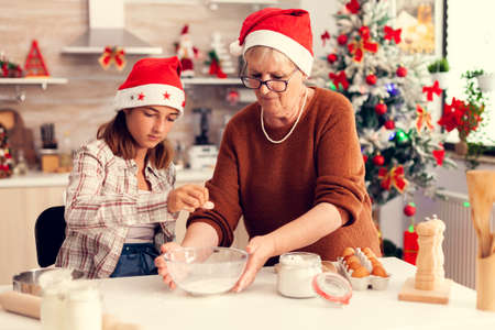 Loving grandmother on christmas day making cookies with niece. Happy cheerful joyfull teenage girl helping senior woman preparing sweet cookies to celebrate winter holidays wearing santa hat.の写真素材