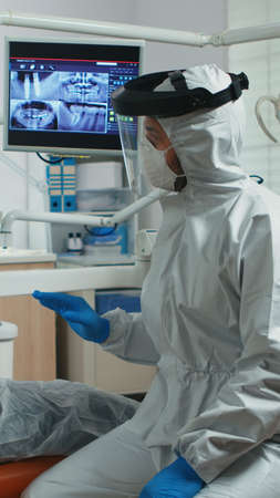 Senior woman showing aching tooth to dentist dressed in coverall during coronavirus epidemic. Assistant and dentist doctor talking with patient wearing protective suit, face shield, mask and glovesの写真素材