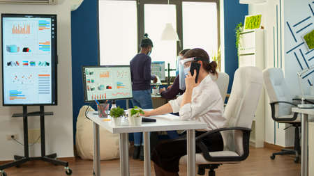 Freelancer with protection mask talking on mobile phone with partner, sitting at desk in office room...