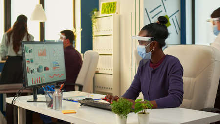 Black woman with protection face masks working on computer in workplace during pandemic. Multiethnic team in new normal business financial office checking reports, analysing datas looking at desktopの写真素材