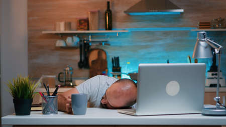 Overworked man sleeping on kitchen desk working remotely late hours at home. Busy exhausted remote employee napping on chair waking up working on laptop using modern technology network overtimeの写真素材