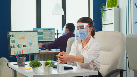 Entrepreneur with protection mask and visor applying sanitizer gel rubbing hands before typing at computer. Businesswoman in new normal workplace disinfecting while colleagues working in backgroundの写真素材