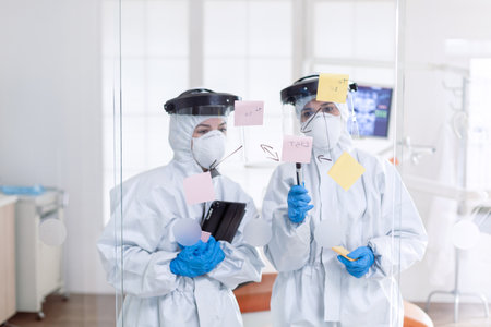 Professional dentists in ppe suit checking schedule during covid-19. Medical team in stomatology office wearing coverall in dental office writing ideas on sticky notes.の写真素材