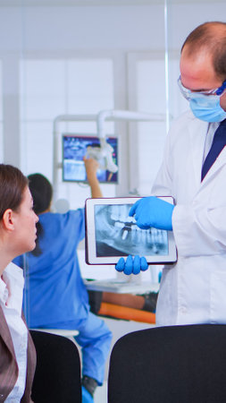 Woman sitting on chair in waiting room of crowded stomatological clinic listening doctor looking on tablet with digital x-ray. Dentist showing to patient teeth radiography using modern gadget.の写真素材
