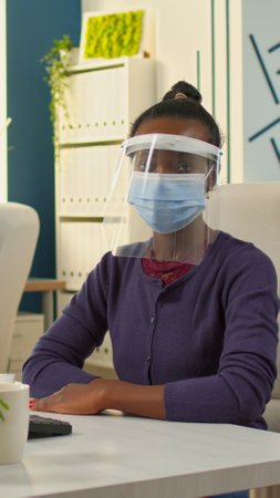 Black employee with visor and protection mask looking serious at camera in new normal office. Multiethnic business team working in financial company respecting social distance during global pandemic.の写真素材