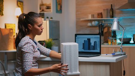 Freelancer architect working in 3D software to elaborate buildings design sitting at kitchen desk at night. Engineer artist creating and studying in office holding scale model, determination, career.の写真素材