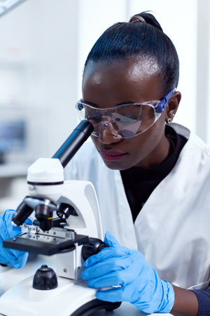 Close up of chemist of african ethnicity doing sample analysis looking through microscope. Black healthcare scientist in biochemistry laboratory wearing sterile equipment.の写真素材