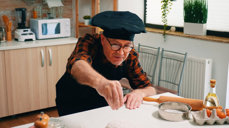 Retired baker man sieving flour on chopping board making dough at home. Elderly senior chef with bonete and uniform sprinkling, sifting, spreading ingredients by hand, baking homemade pizza and breadの写真素材