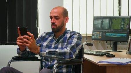 Disabled photo designer sitting in wheelchair searching on internet using smartphone texting,...