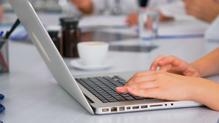 Close up of assistant using laptop, writing meeting informations while doctors discussing in background during medical conference sitting at desk in hospital office. Profesional team brainstormingの写真素材