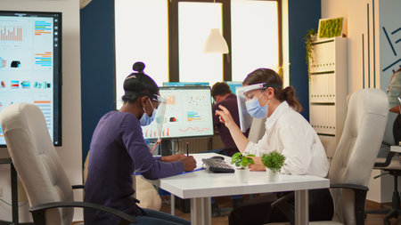 Manager talking with african employee about company statistics wearing protection face mask and visor against coronavirus sitting at desk respecting social distance. New normal business office.の写真素材