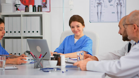Nurse writing on laptop during conference and looking at camera smiling sitting on desk in hospital office, brainstorming room. Specialist woman in lab coat typing on notepad in medical clinic.の写真素材