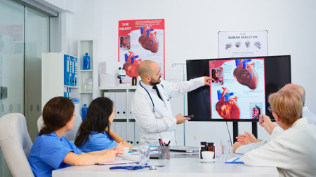 Experienced man doctor analysing heart issues image together with cvalified colleagues in meeting room, pointing on monitor. Doctors discussing diagnosis about treatment of patientsの写真素材