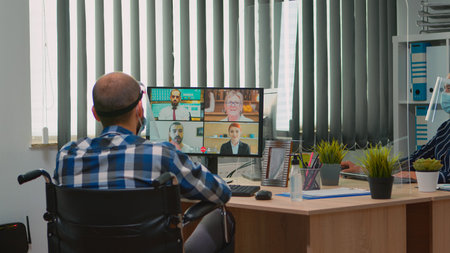 Paralysed manager in wheelchair talking with mask and visor during videocall having online conference in business office. Immobilized freelancer working in financial company respecting social distanceの写真素材